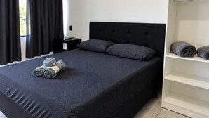 A bedroom with a bed, pillows, a nightstand, a shelf with towels, and a window with curtains. - Pousada Tio Tonho (Penha)