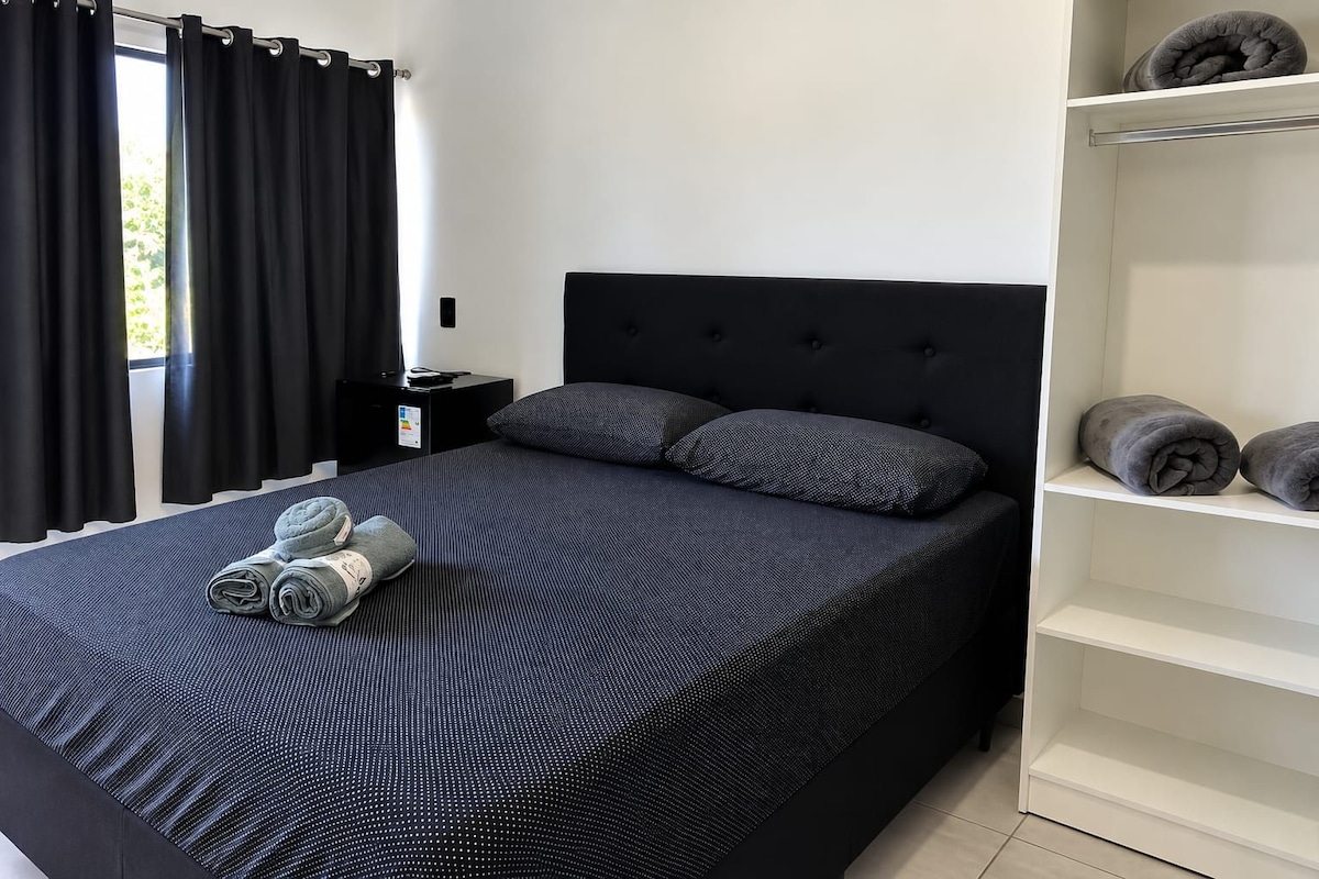 Ein Schlafzimmer mit einem Bett, Kissen, einem Nachttisch, einem Regal mit Handtüchern und einem Fenster mit Vorhängen.