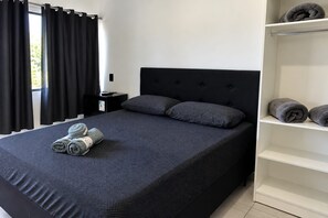 Ein Schlafzimmer mit einem Bett, Kissen, einem Nachttisch, einem Regal mit Handtüchern und einem Fenster mit Vorhängen.