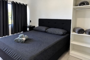 Un dormitorio con cama, almohadas, una mesita de noche, un estante con toallas y una ventana con cortinas.