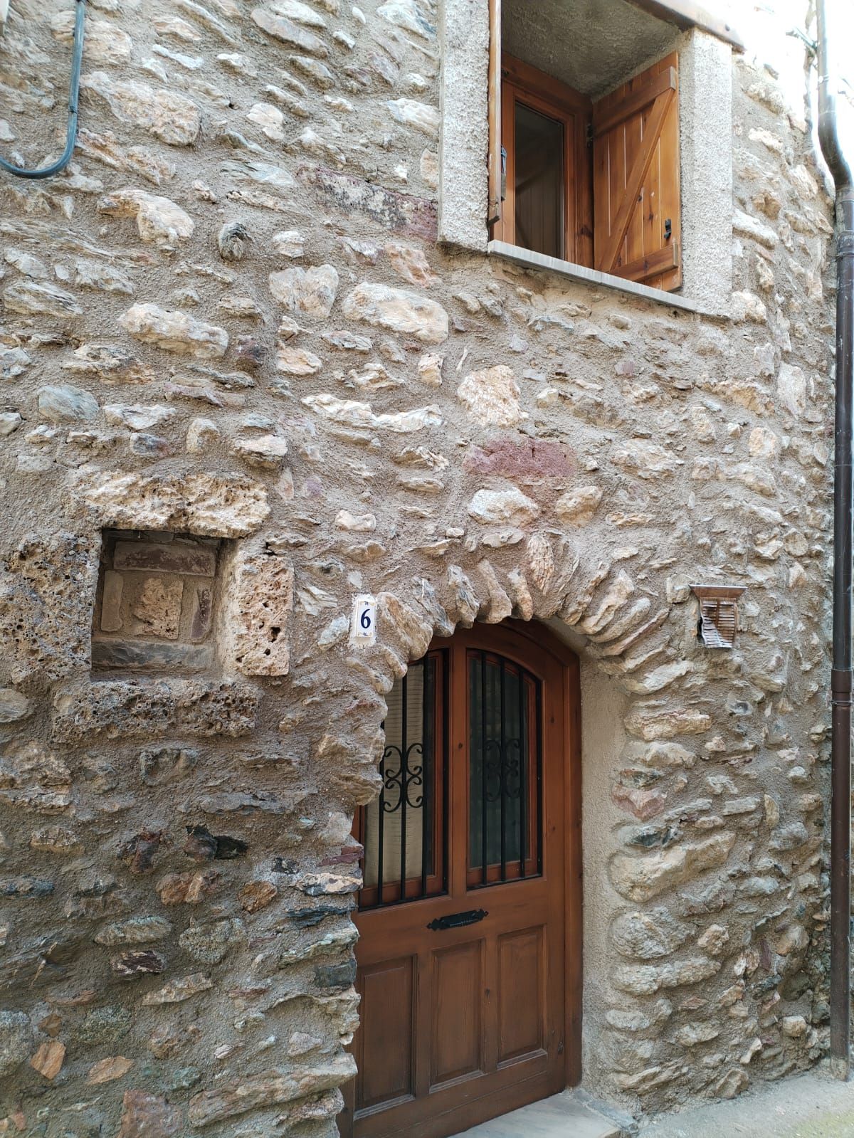 A stone building with a wooden door and window, a small window with shutters, and a number plaque.