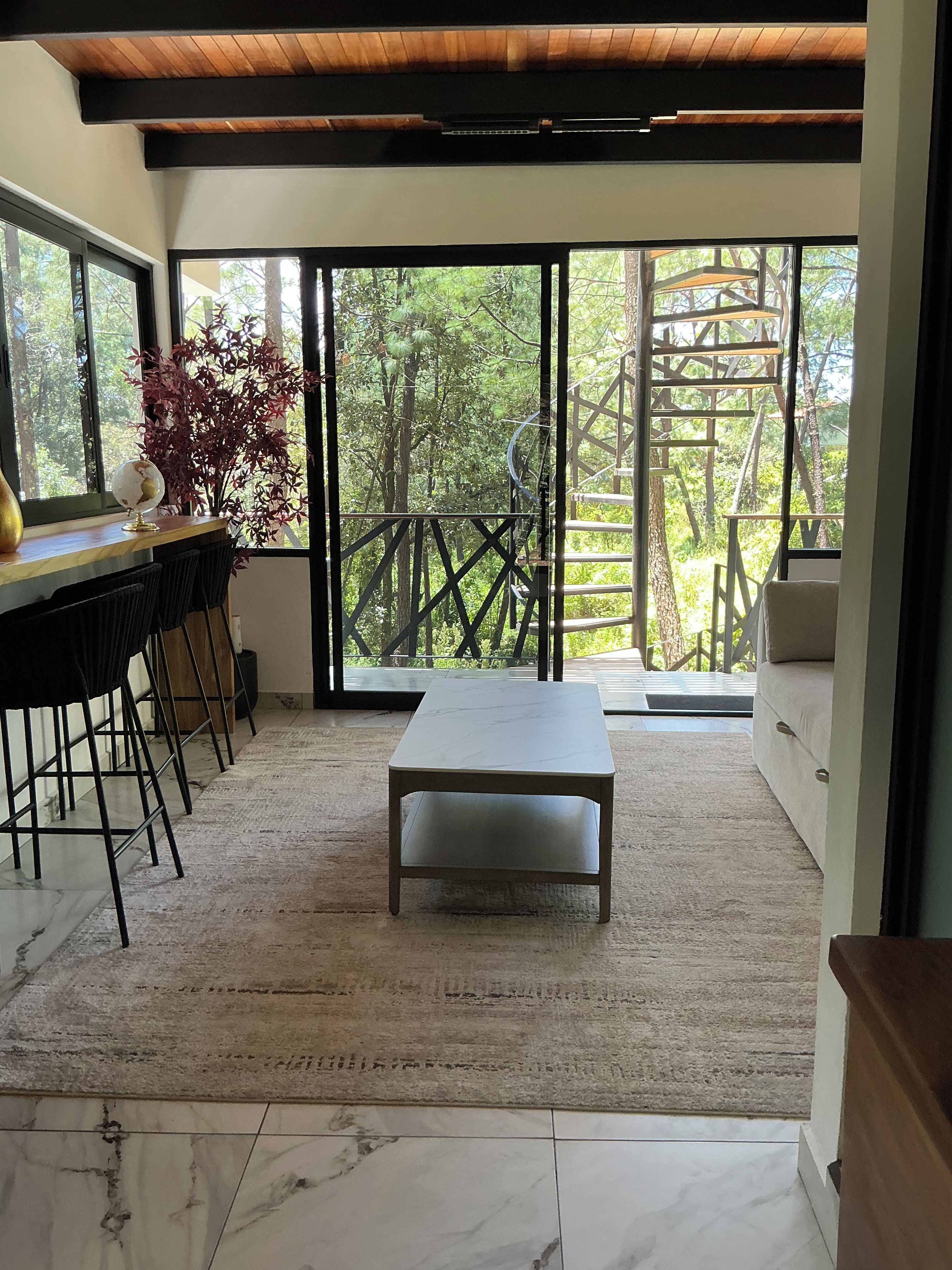Un intérieur moderne avec un sol en marbre, un plafond en bois, un coin repas comprenant des chaises et une table, et une grande porte-fenêtre coulissante donnant sur un balcon avec vue sur les arbres.