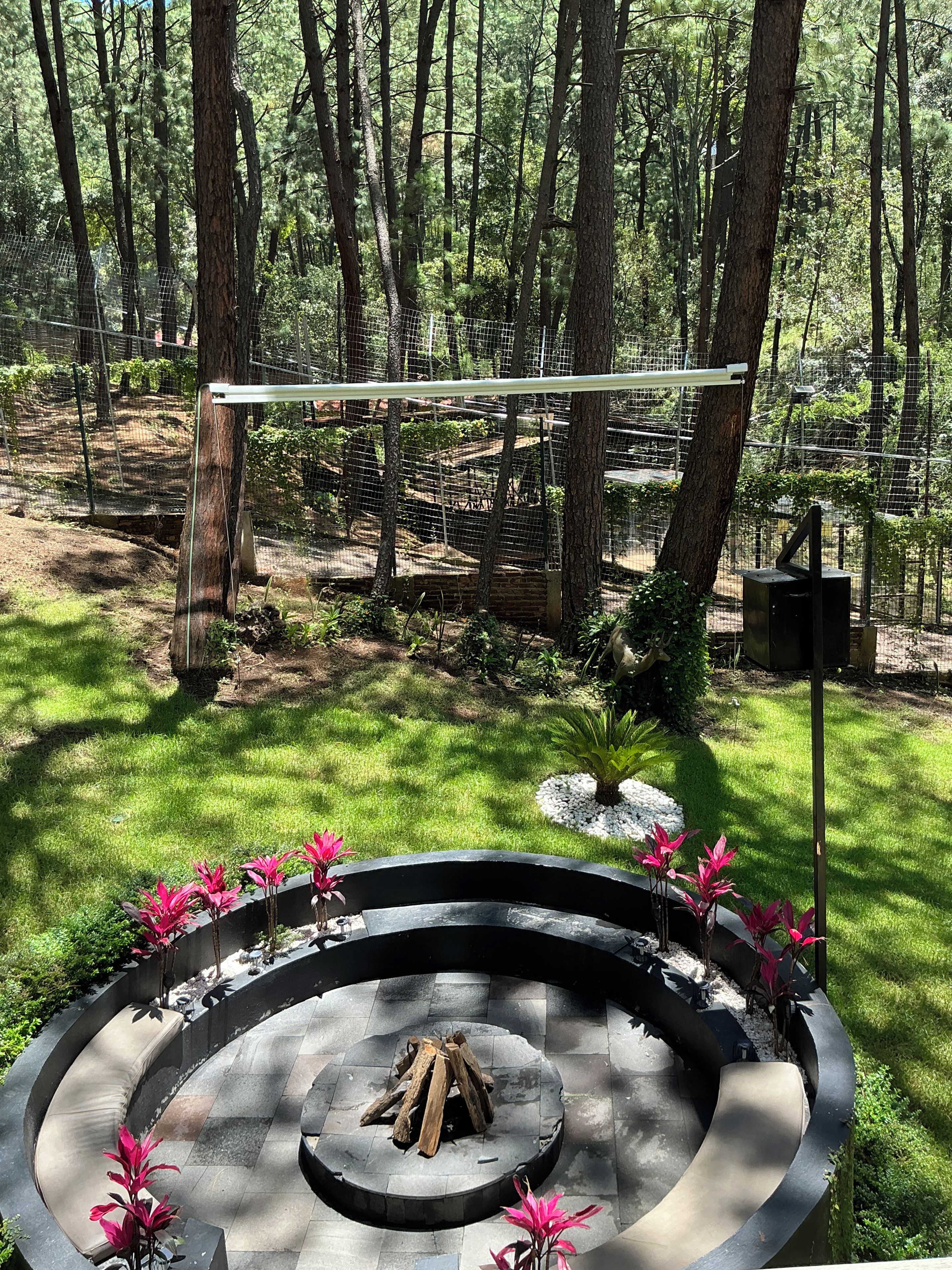 A circular stone fire pit with logs, surrounded by a low stone wall, in a forested outdoor area with a metal fence and a wooden structure in the background.