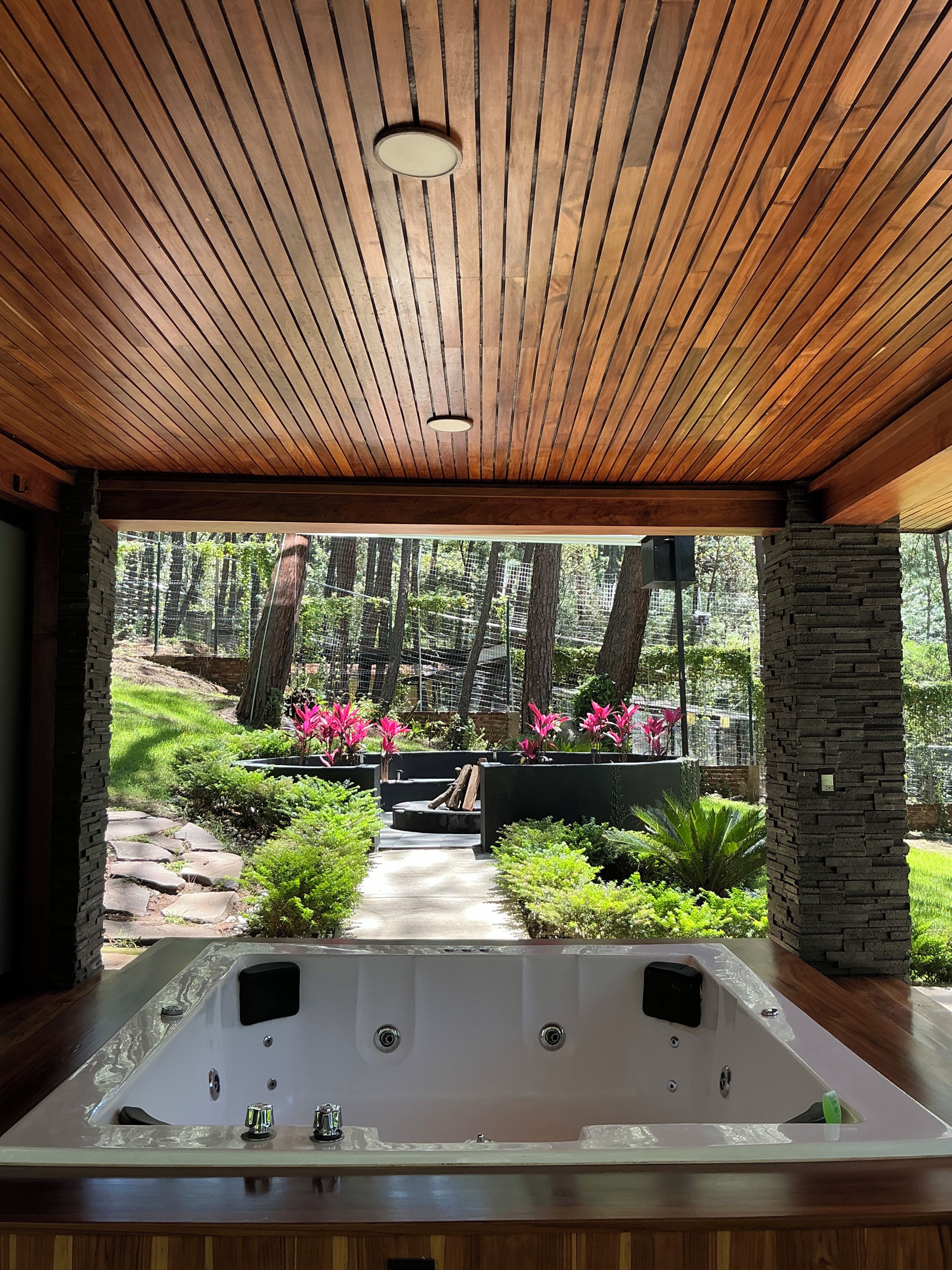 A spa area with a wooden ceiling, a hot tub, and a view of a garden with trees and plants.