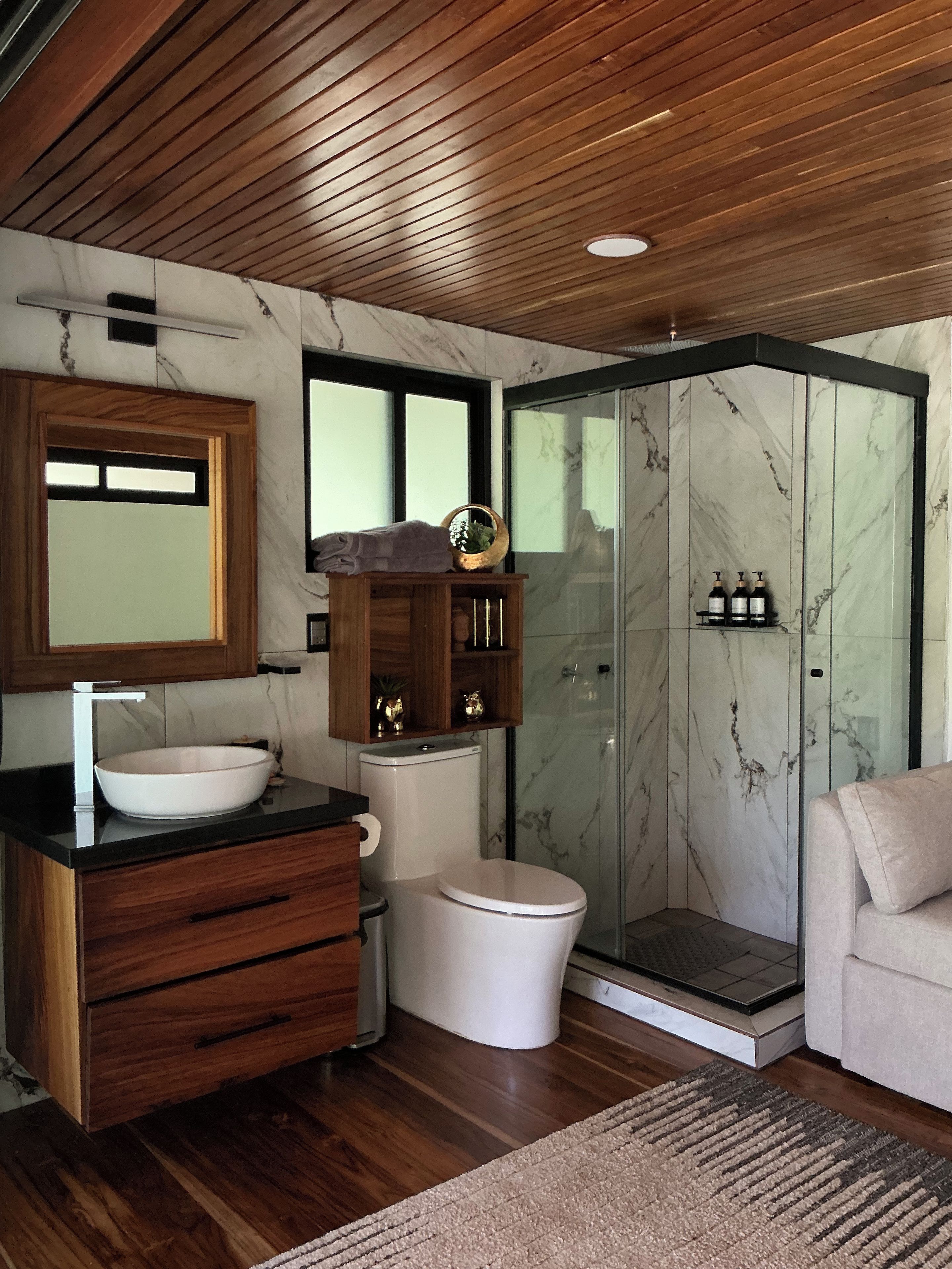 A modern bathroom with a wooden vanity, a large mirror, a white toilet, a glass shower enclosure, and a beige sofa.