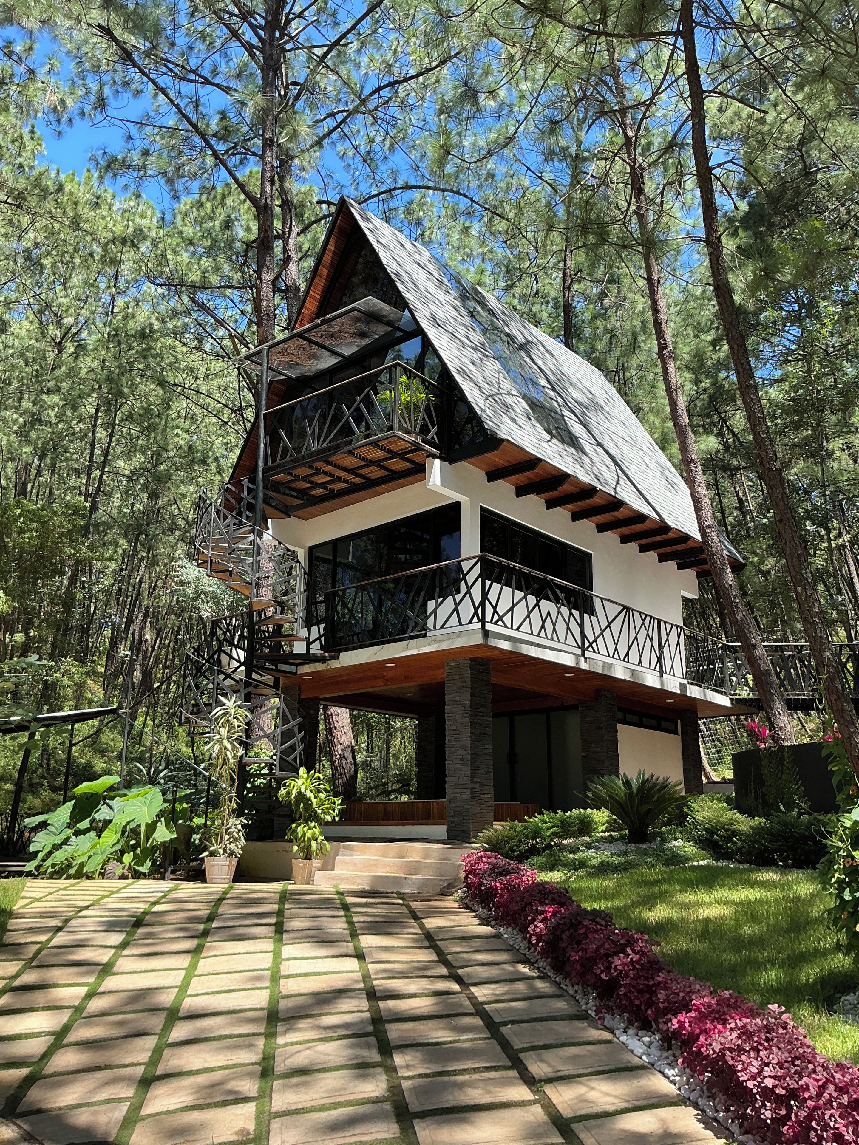 Une cabane à deux étages dotée d’une terrasse en bois et d’un balcon, entourée d’une forêt.
