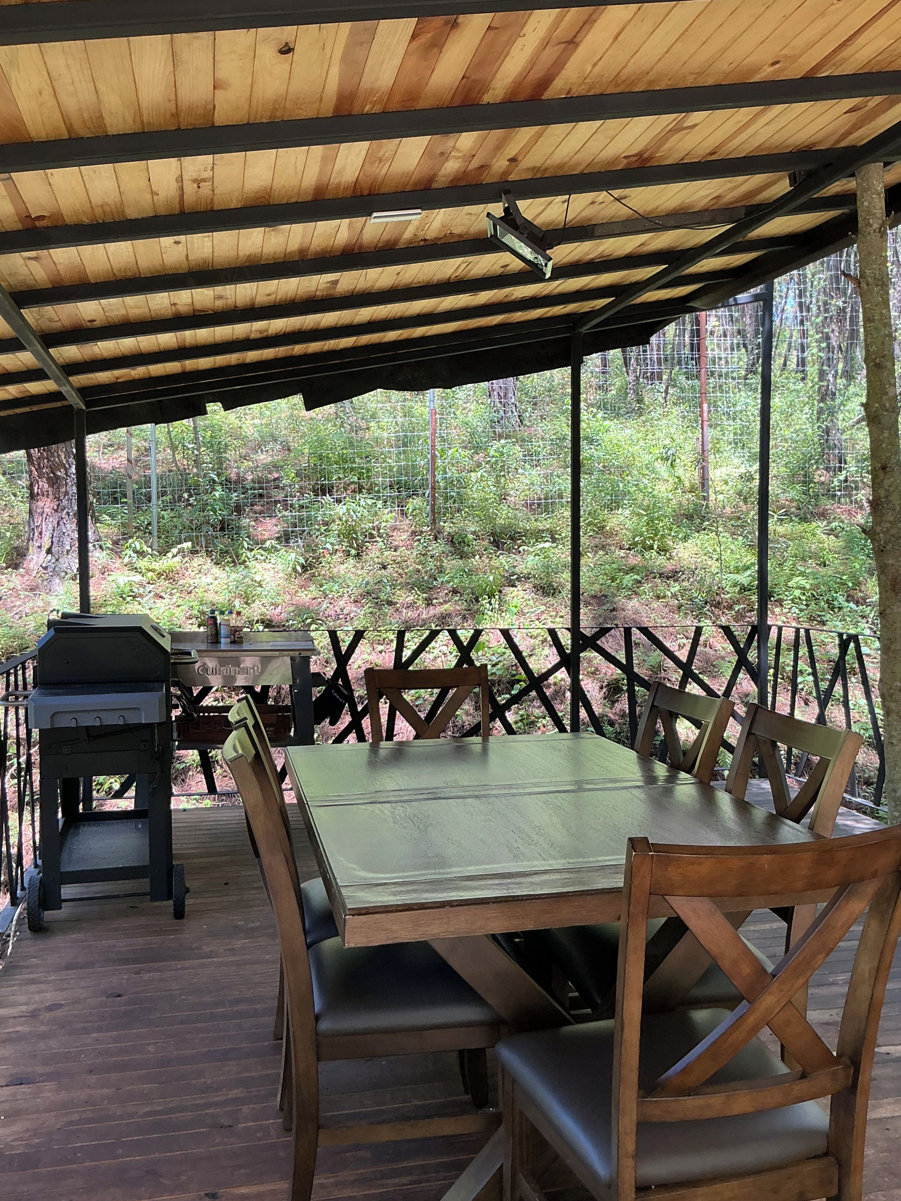 Un espace de restauration extérieur abrité, avec une table et des chaises, un barbecue et une vue sur une zone boisée.
