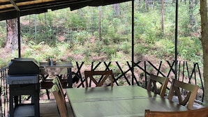 Un espace de restauration extérieur abrité, avec une table et des chaises, un barbecue et une vue sur une zone boisée.