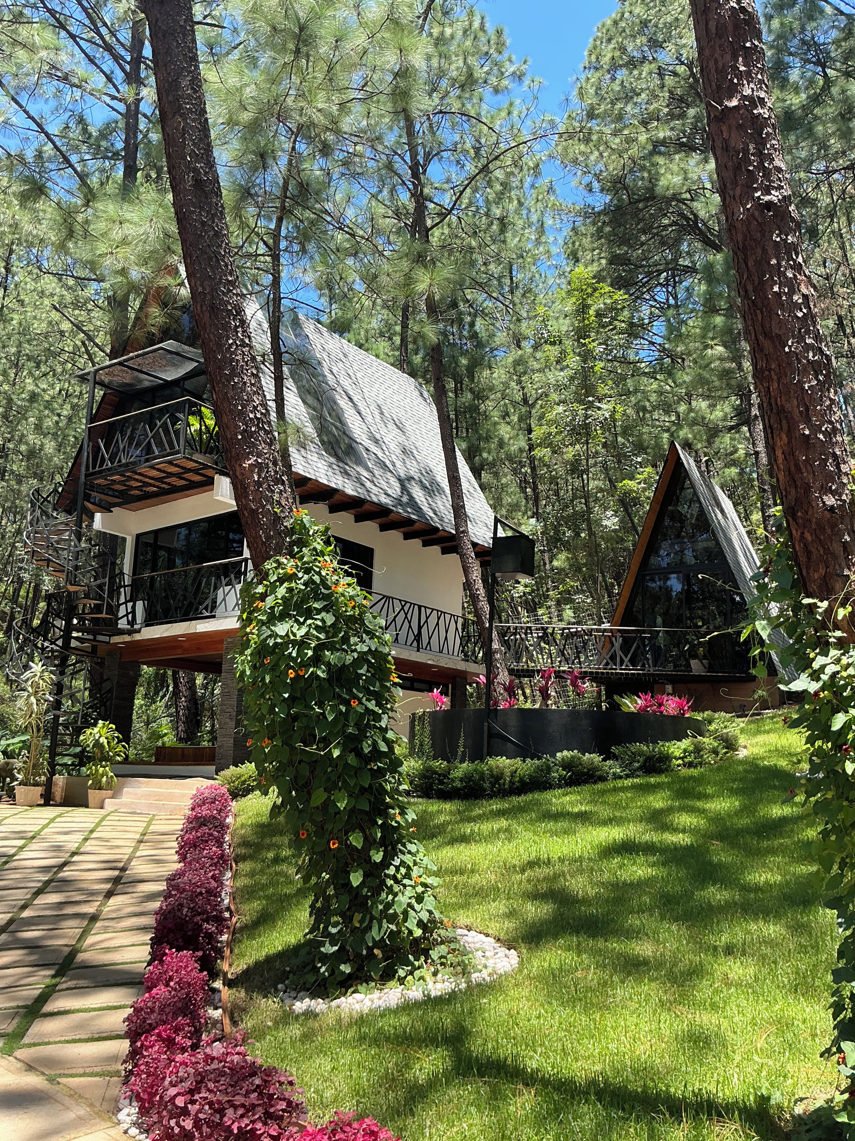 A two-story cabin with a wooden deck and a glass enclosure, surrounded by a well-maintained garden and tall pine trees.