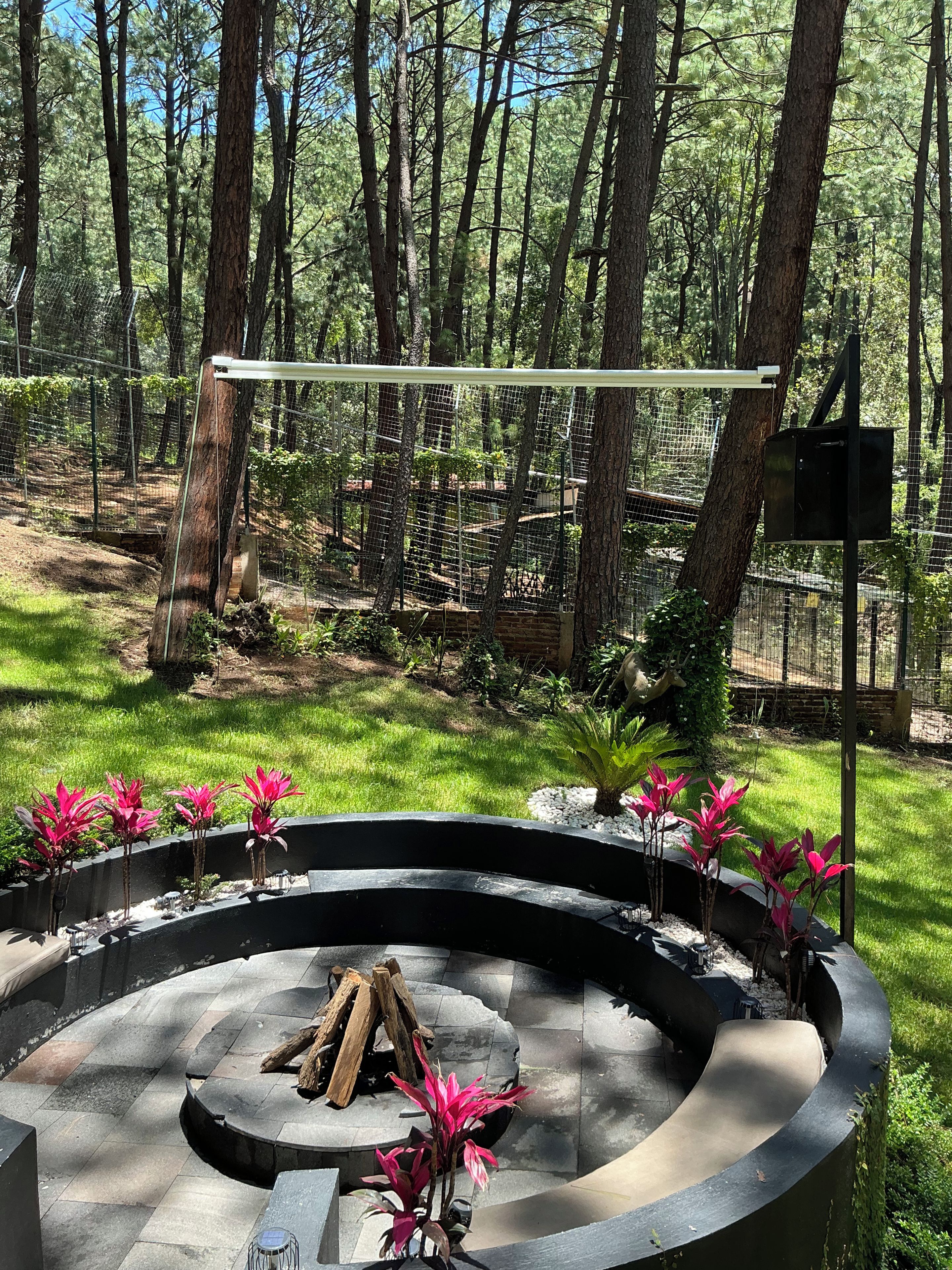 Un área de estar al aire libre con forma circular, que incluye un brasero, rodeada por una valla metálica y árboles.