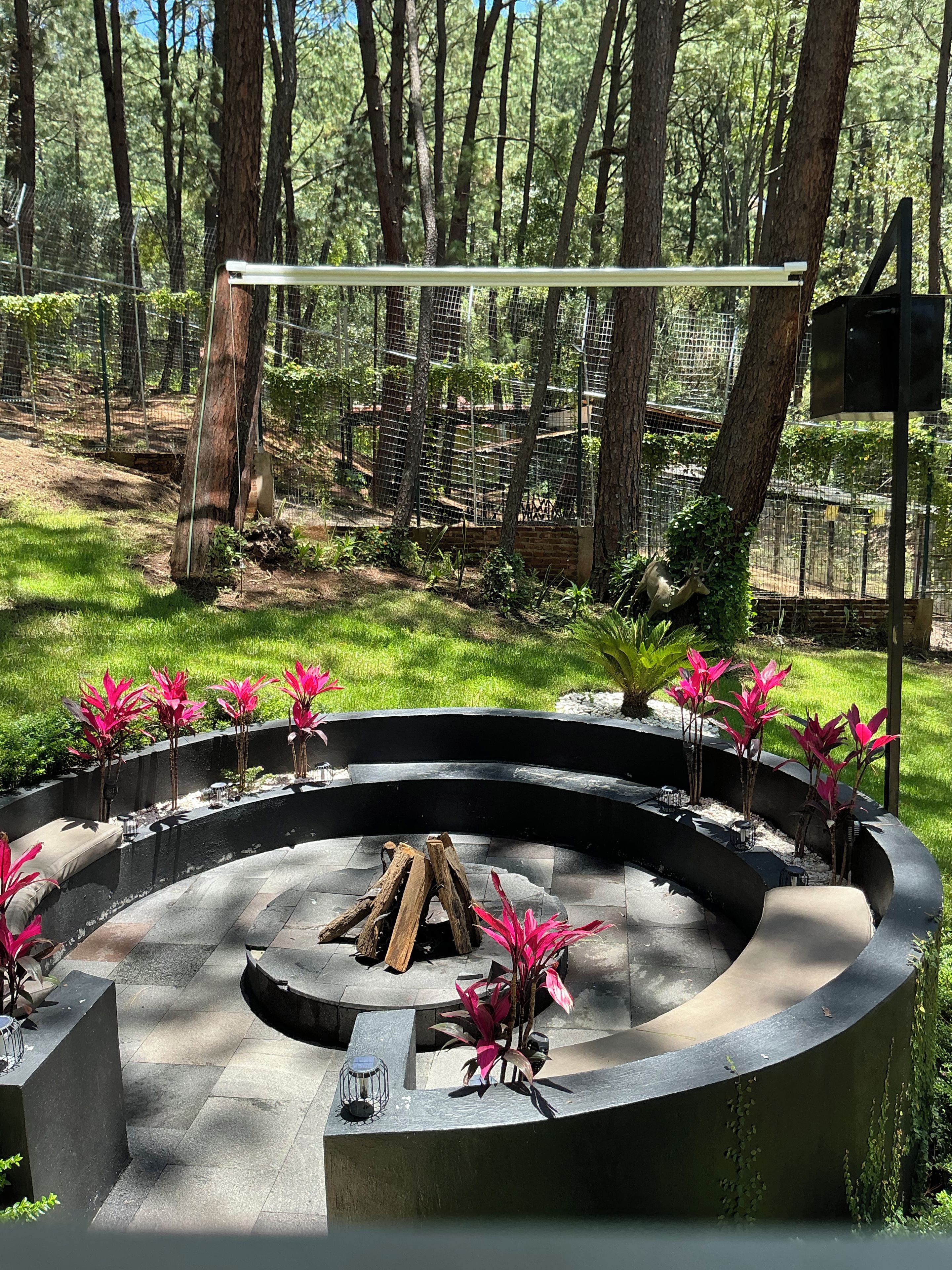 Un foyer circulaire en pierre, avec des bûches, entouré d’une clôture métallique, situé dans une zone boisée avec des arbres et de l’herbe.