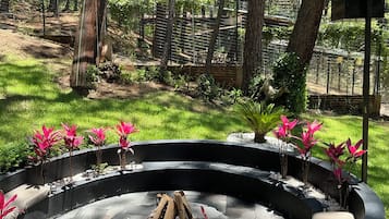 Un foyer circulaire en pierre, avec des bûches, entouré d’une clôture métallique, situé dans une zone boisée avec des arbres et de l’herbe.
