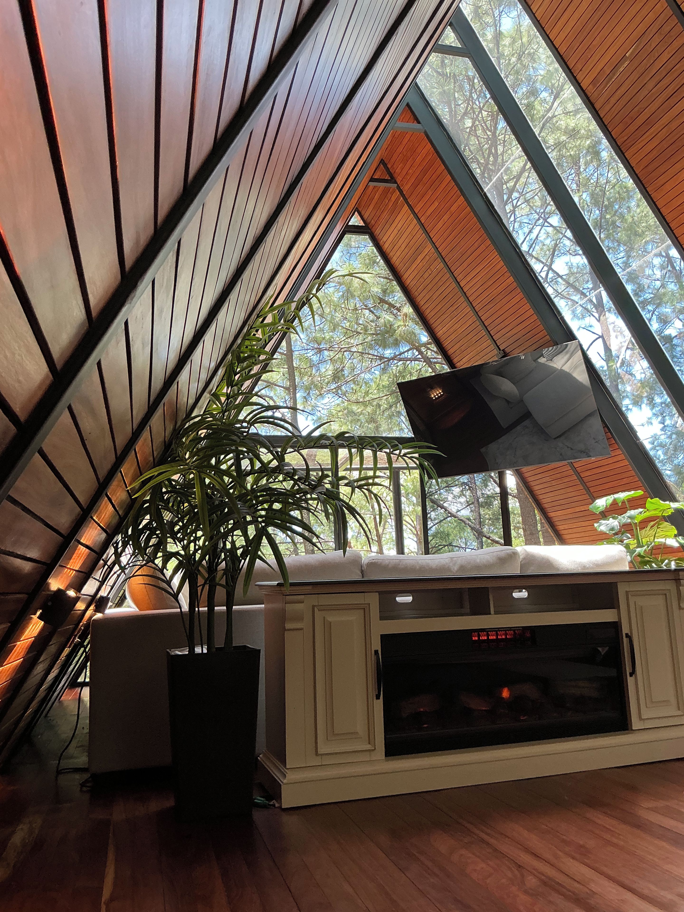 A room with a sloped ceiling, a television mounted on the wall, a fireplace, and a large window with a view of trees.