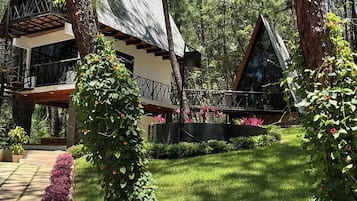 Une cabane dotée d’un toit en pente, entourée d’un jardin bien entretenu où poussent diverses plantes, et accessible par un sentier en pierre.