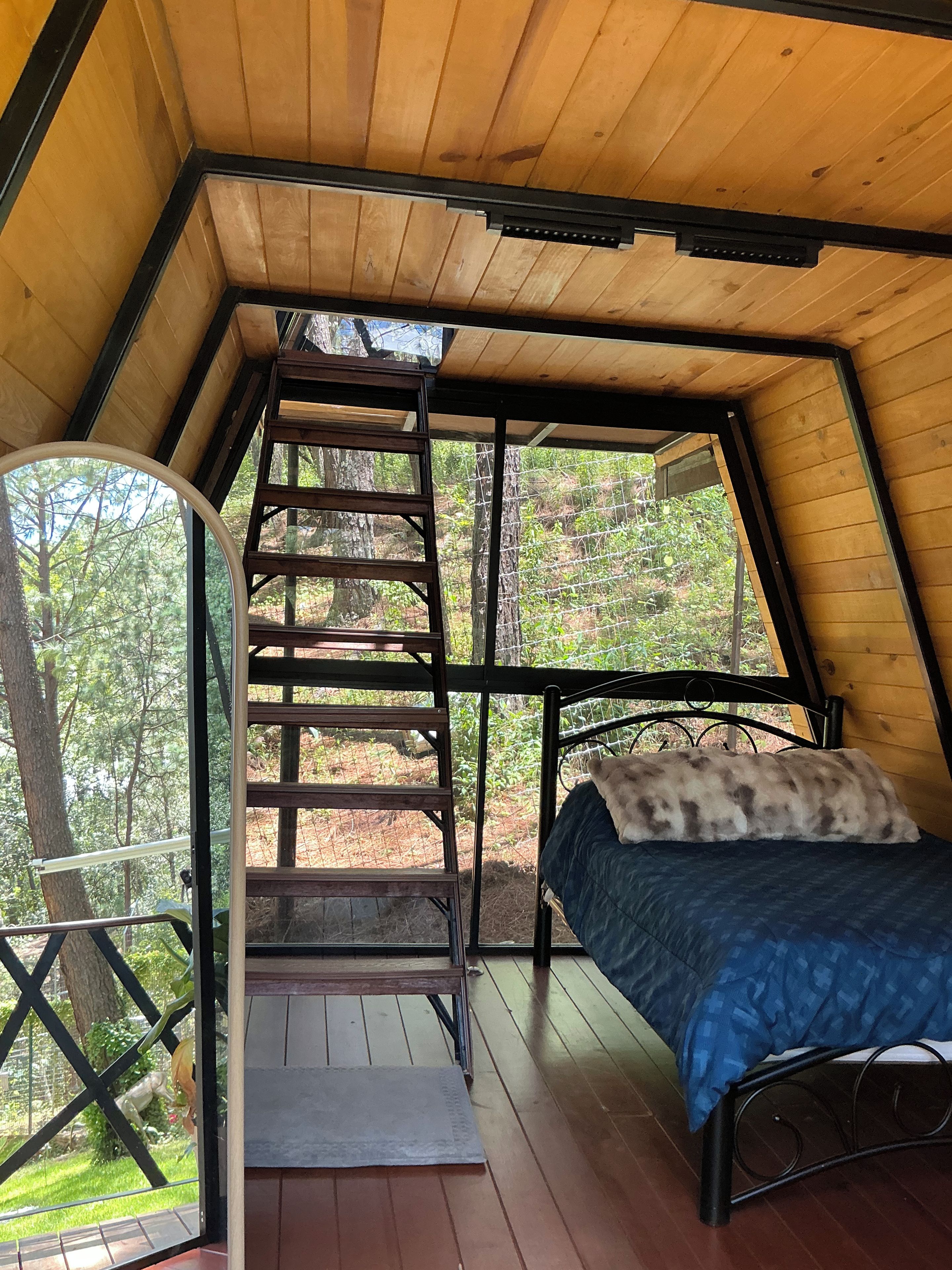 Interior de madera con una cama, una escalera y un ventanal con vistas a un bosque.