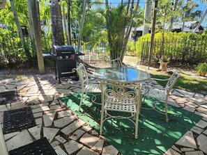 Eine Terrasse im Freien mit einem runden Glastisch und weißen Metallstühlen, umgeben von einem steinbelagten Boden und künstlichem Rasen.