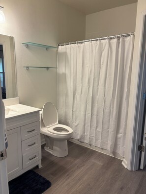 A bathroom with a toilet, a glass shelf, a shower curtain, and a dark blue rug. - 2-bedroom condo on first floor in peaceful Seminole, FL-5 min from Maderia Beach (Seminole)