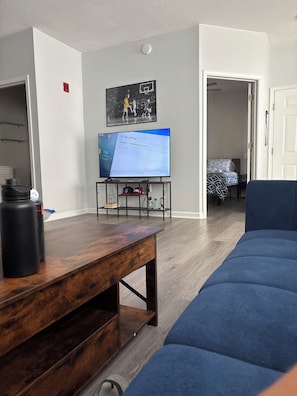 Ein Wohnzimmer mit Couchtisch aus Holz, einem blauen Sofa, einem Fernseher auf einem Schrank und einer Tür zu einem weiteren Raum.