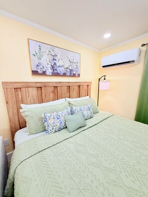 Ein Schlafzimmer mit einem hölzernen Kopfende, einem Bett mit einer gemusterten grünen Bettdecke, floralen Kissen, einer Wand-Klimaanlage und einem Bild mit Vasen.