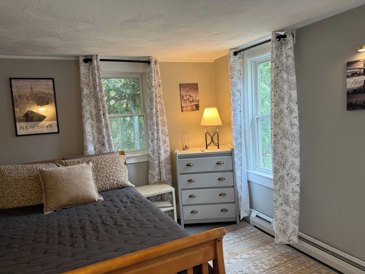 Une chambre à coucher avec un lit, une commode, une lampe et deux fenêtres avec des rideaux.