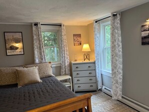 Un dormitorio con cama, cómoda, lámpara y dos ventanas con cortinas.
