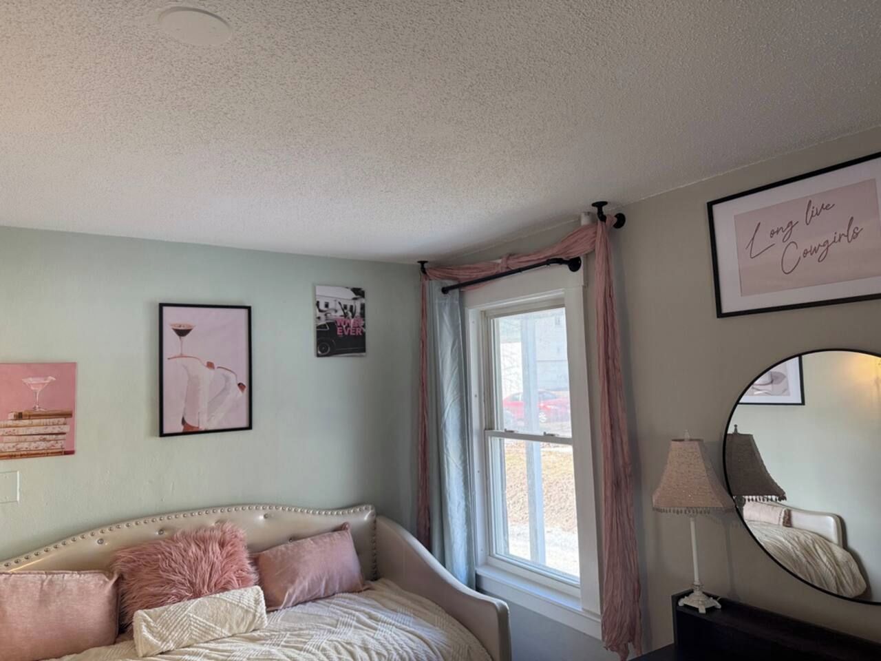 A bedroom with a bed, a window with curtains, a wall-mounted mirror, and framed pictures.