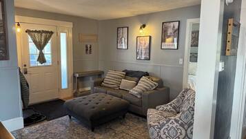 A living room with a sofa, ottoman, patterned rug, and wall-mounted artwork.
