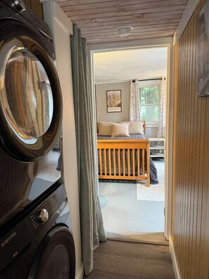 A laundry area with a washing machine and dryer, leading to a bedroom with a bed, pillows, and a window with curtains.