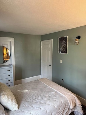 Quarto com cama, cômoda, porta e luminária fixada na parede.
