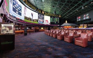 Un'ampia area interna con file di poltrone in pelle, un grande schermo che mostra contenuti sportivi e un'area bar tipo Clubhouse.