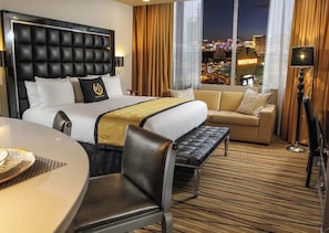 Una camera d'hotel con un letto grande, un divano, un tavolo da pranzo e vista sulla città illuminata di notte.