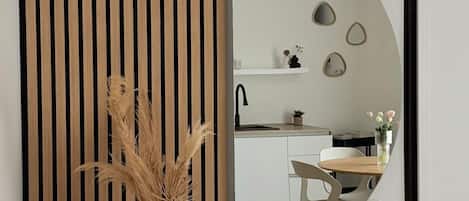 Un interior moderno con un separador de listones de madera, un jarrón con plantas secas y un espejo que refleja una cocina.