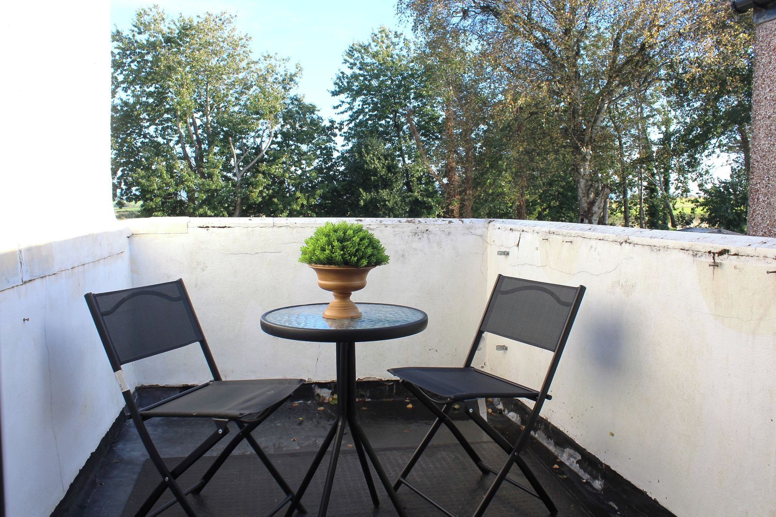 A small outdoor seating area with two chairs and a table, surrounded by a white wall and trees in the background.