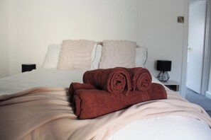 Una cama bien hecha con toallas enrolladas, una mesita de noche con una lámpara y una puerta al fondo.