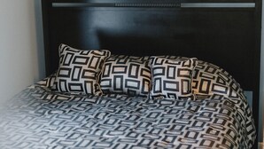 Una cama con una colcha y almohadas de estampado geométrico, un cabecero oscuro y una pared lisa.