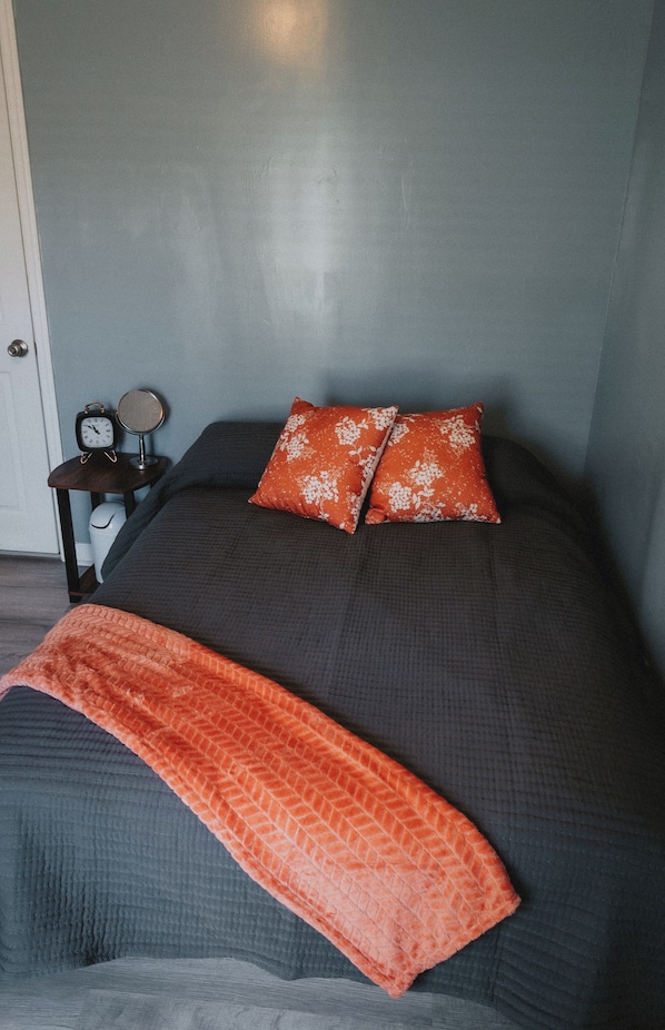 A bed with a dark grey bedspread, two orange pillows with white floral patterns, and a matching orange throw blanket.