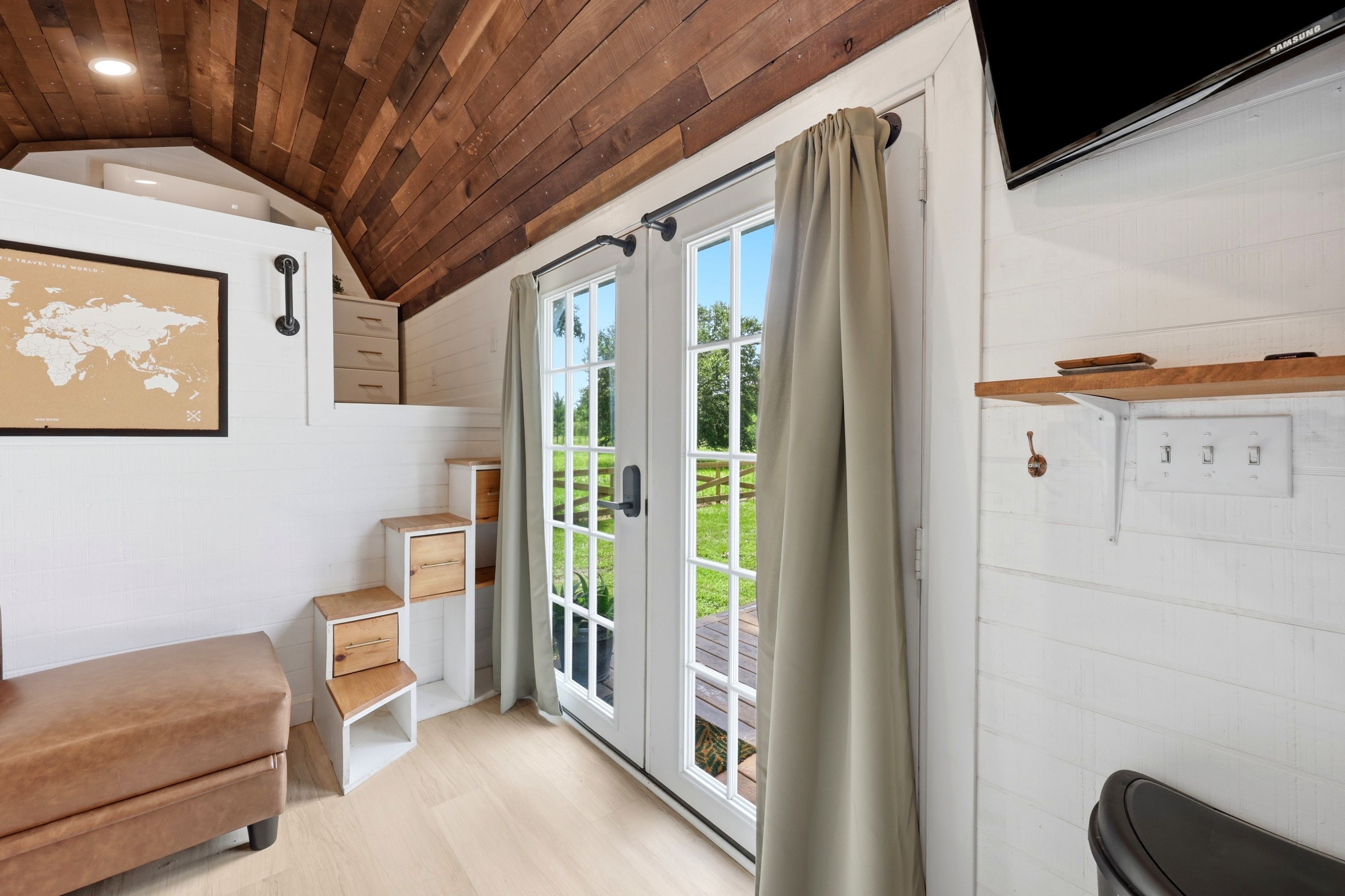 Una habitación con techo de madera, un ventanal con cortinas, un televisor de pantalla plana en la pared y una pequeña zona de estar con un banco y cajones.