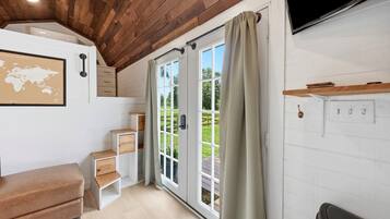 Una habitación con techo de madera, un ventanal con cortinas, un televisor de pantalla plana en la pared y una pequeña zona de estar con un banco y cajones.