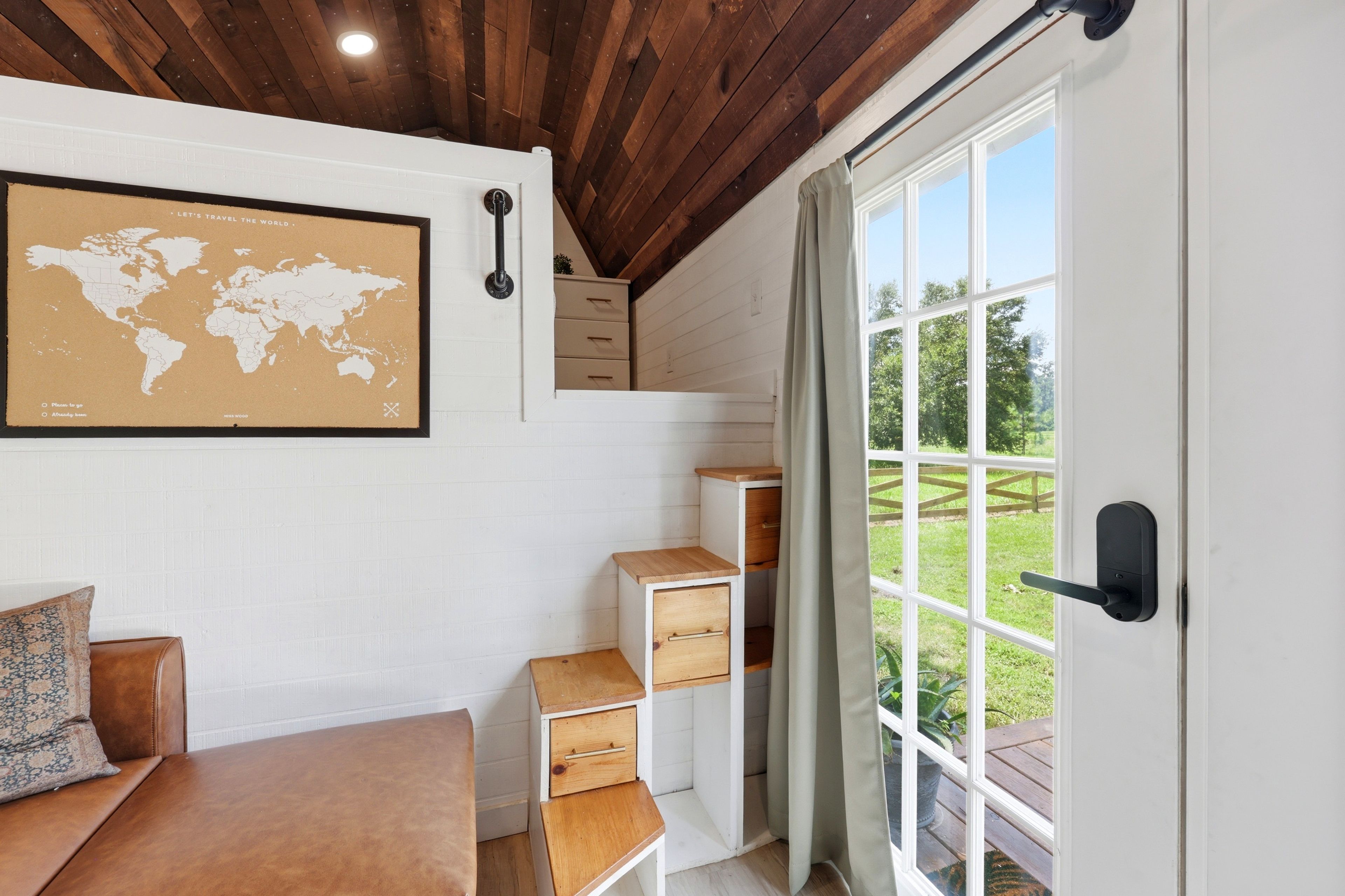 A room with a wooden ceiling, a world map on the wall, a wooden desk with drawers, a bench, and a view of a garden through a glass door.