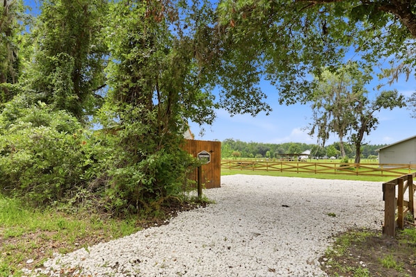 A gravel driveway leading to a wooden gate with a sign, surrounded by lush greenery and a clear sky.