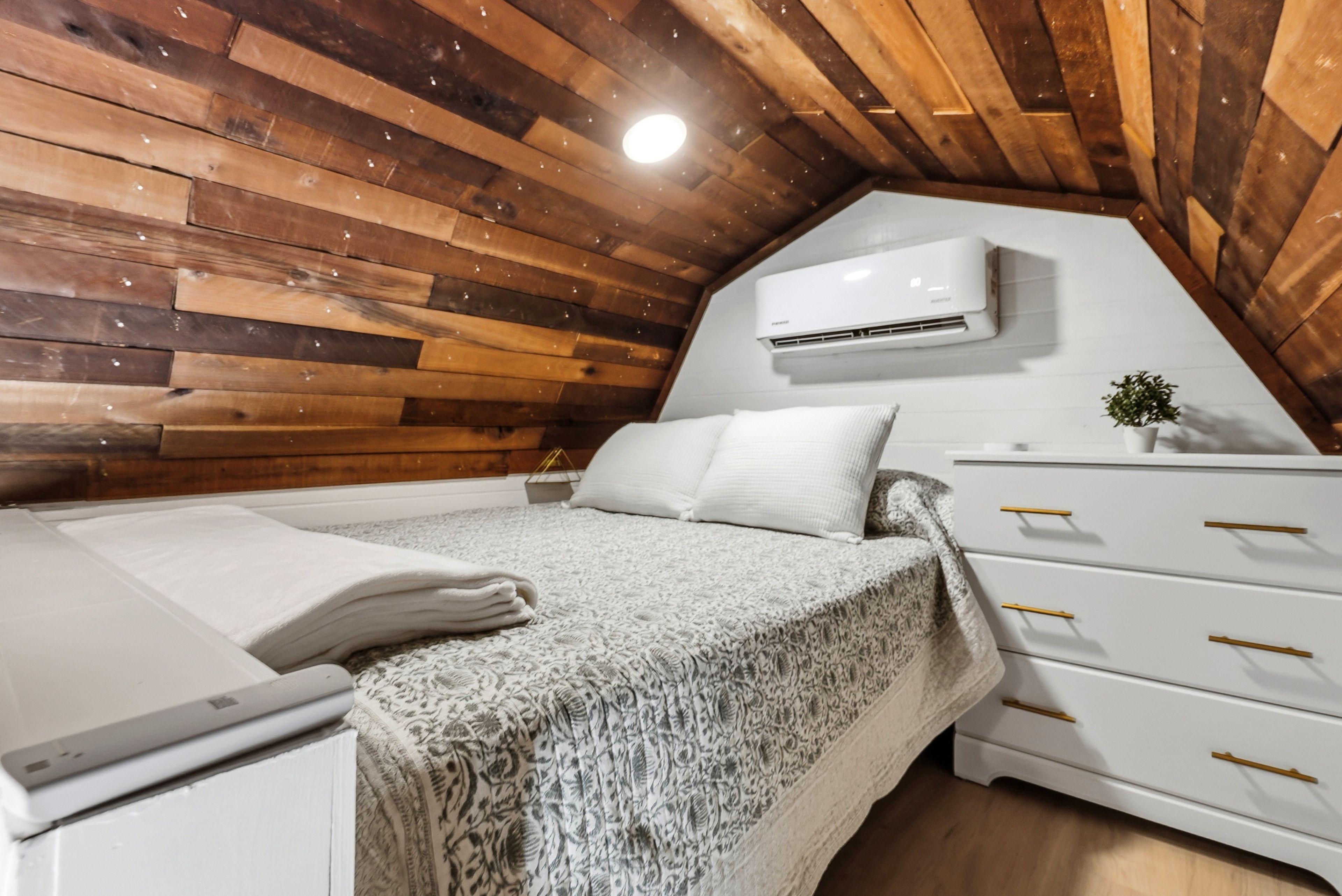 Een slaapkamer met een schuine kap, houten lambrisering, een bed met witte lakens, een witte ladekast met gouden handgrepen en een airconditioning.