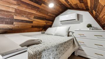 Een slaapkamer met een schuine kap, houten lambrisering, een bed met witte lakens, een witte ladekast met gouden handgrepen en een airconditioning.