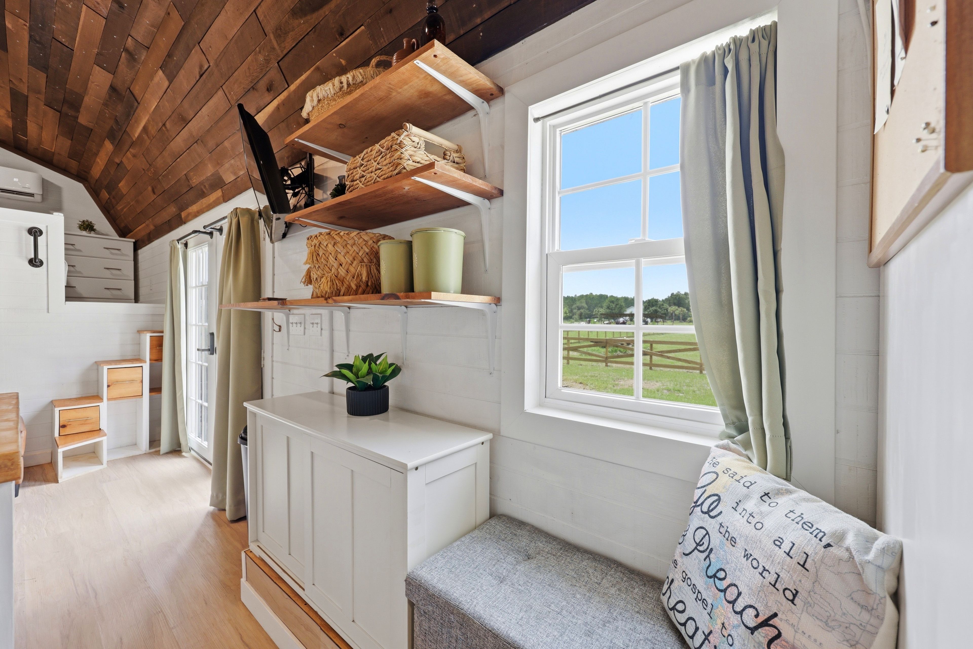 Una habitación acogedora con techo de madera, paredes blancas, una ventana con cortinas, un banco y una estantería con cestas y recipientes.