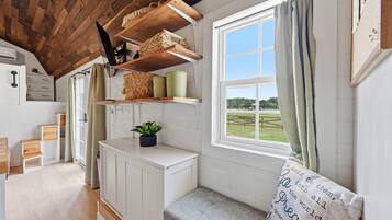 Una habitación acogedora con techo de madera, paredes blancas, una ventana con cortinas, un banco y una estantería con cestas y recipientes.