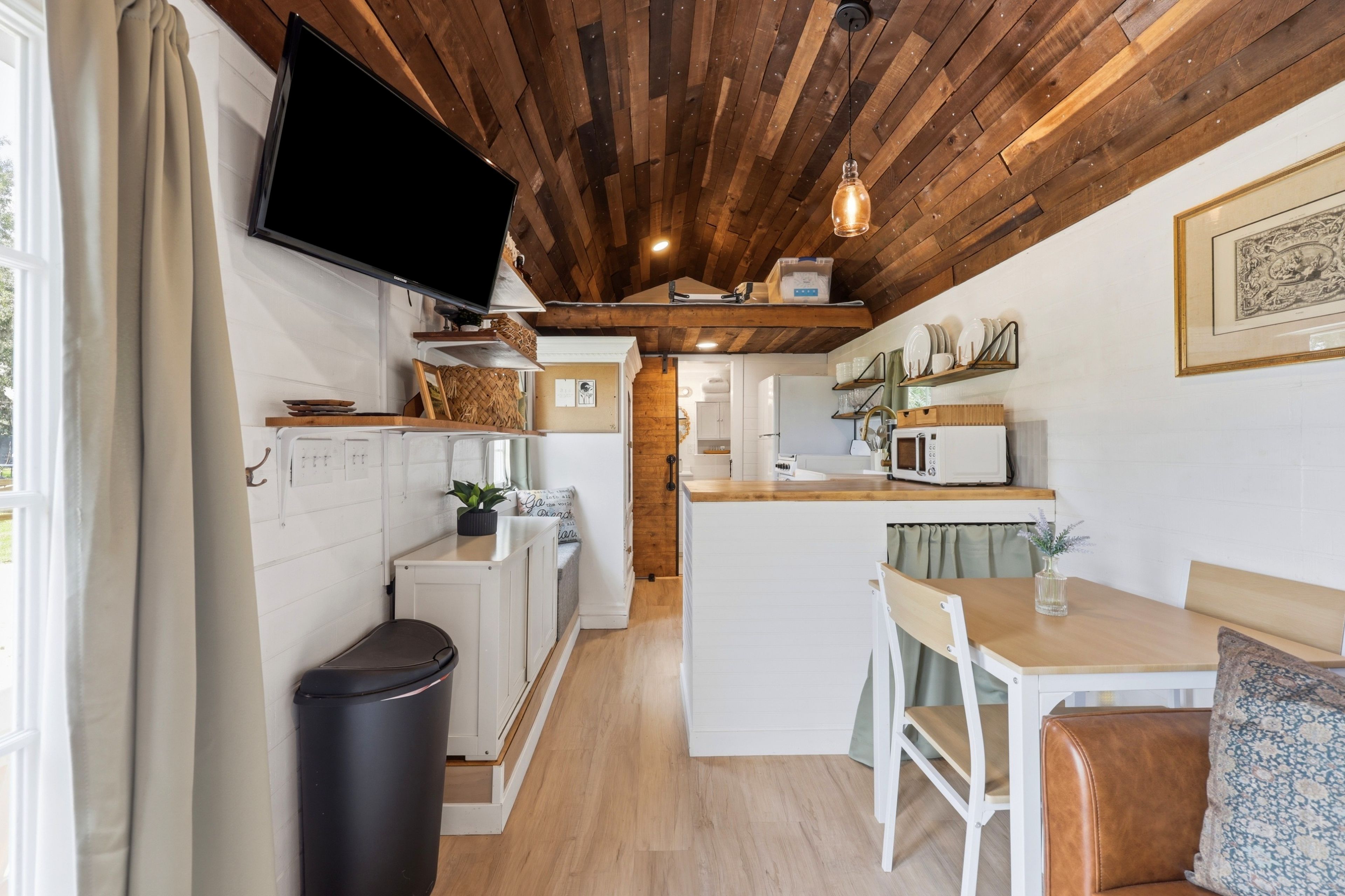 A compact kitchen with a wooden ceiling, a television mounted above the counter, and a dining area with a table and chairs.
