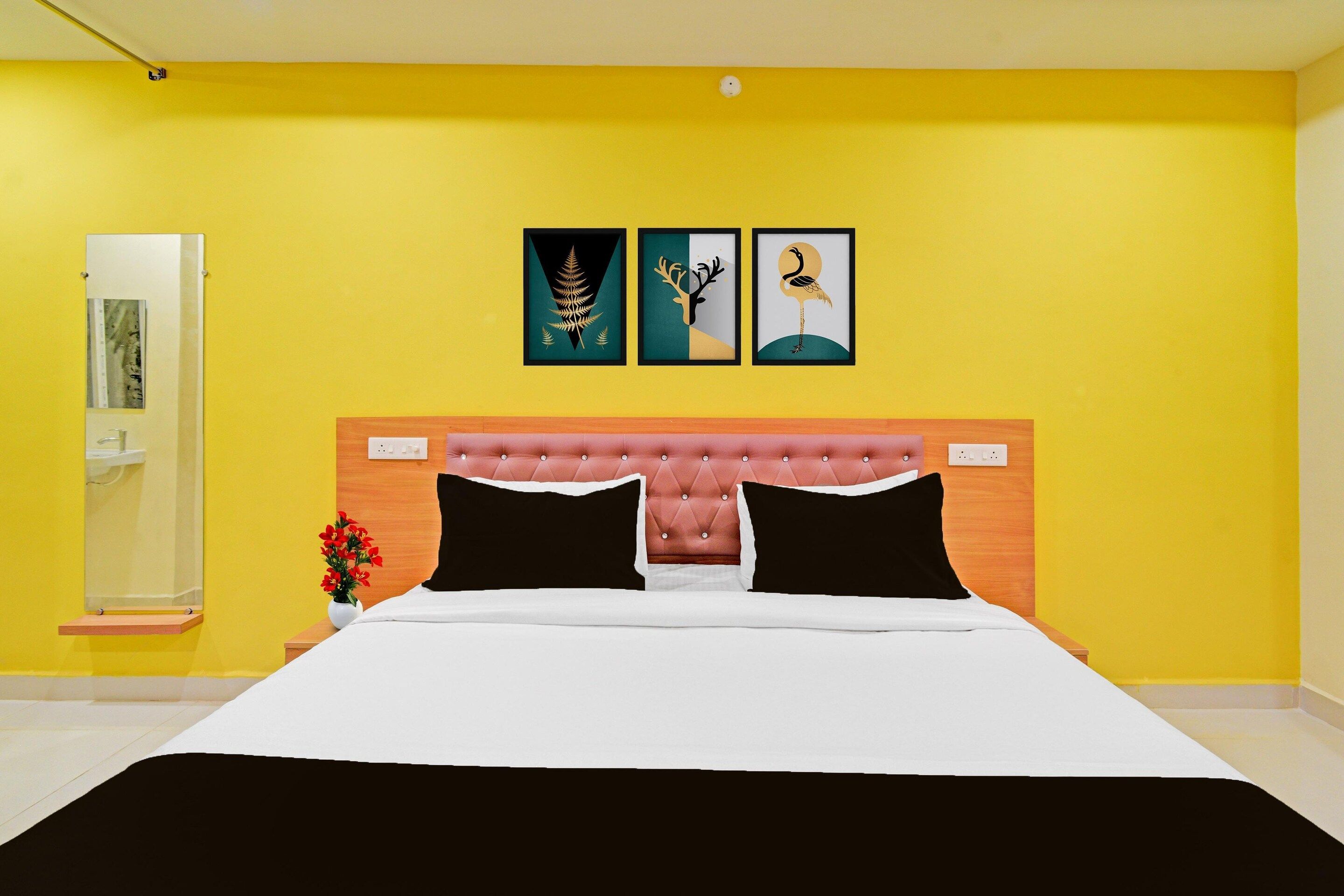 A hotel room with a yellow wall, a tufted headboard, a bed with white and black bedding, and three framed artworks above the bed.