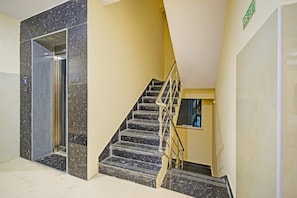 A staircase with a handrail, a fire exit sign, and a glass elevator door. - Classic Gachibowli Bus Terminal (Hyderabad)