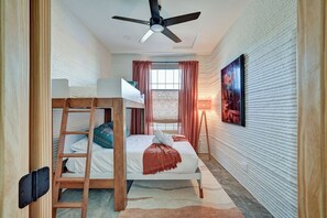 Een kamer met stapelbedden, een plafondventilator, een raam met gordijnen en een schilderij aan de muur.