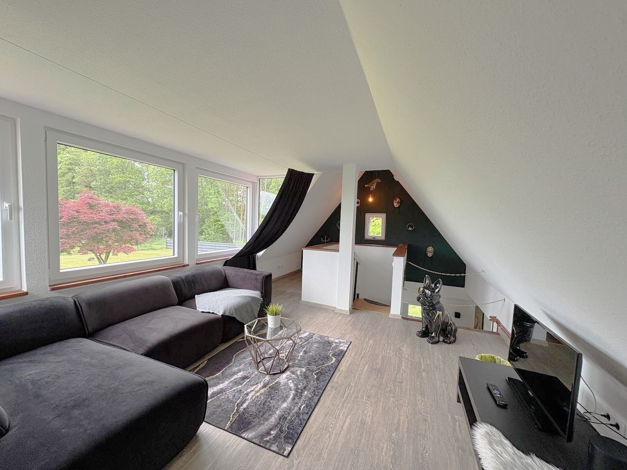 A modern loft bedroom with a large window, a built-in desk, and a seating area with a sofa.