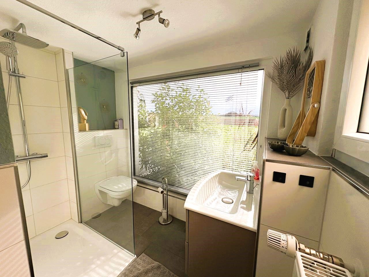 Ein modernes Badezimmer mit Glasdusche, Toilette, Waschbecken und einem Fenster mit Jalousien.